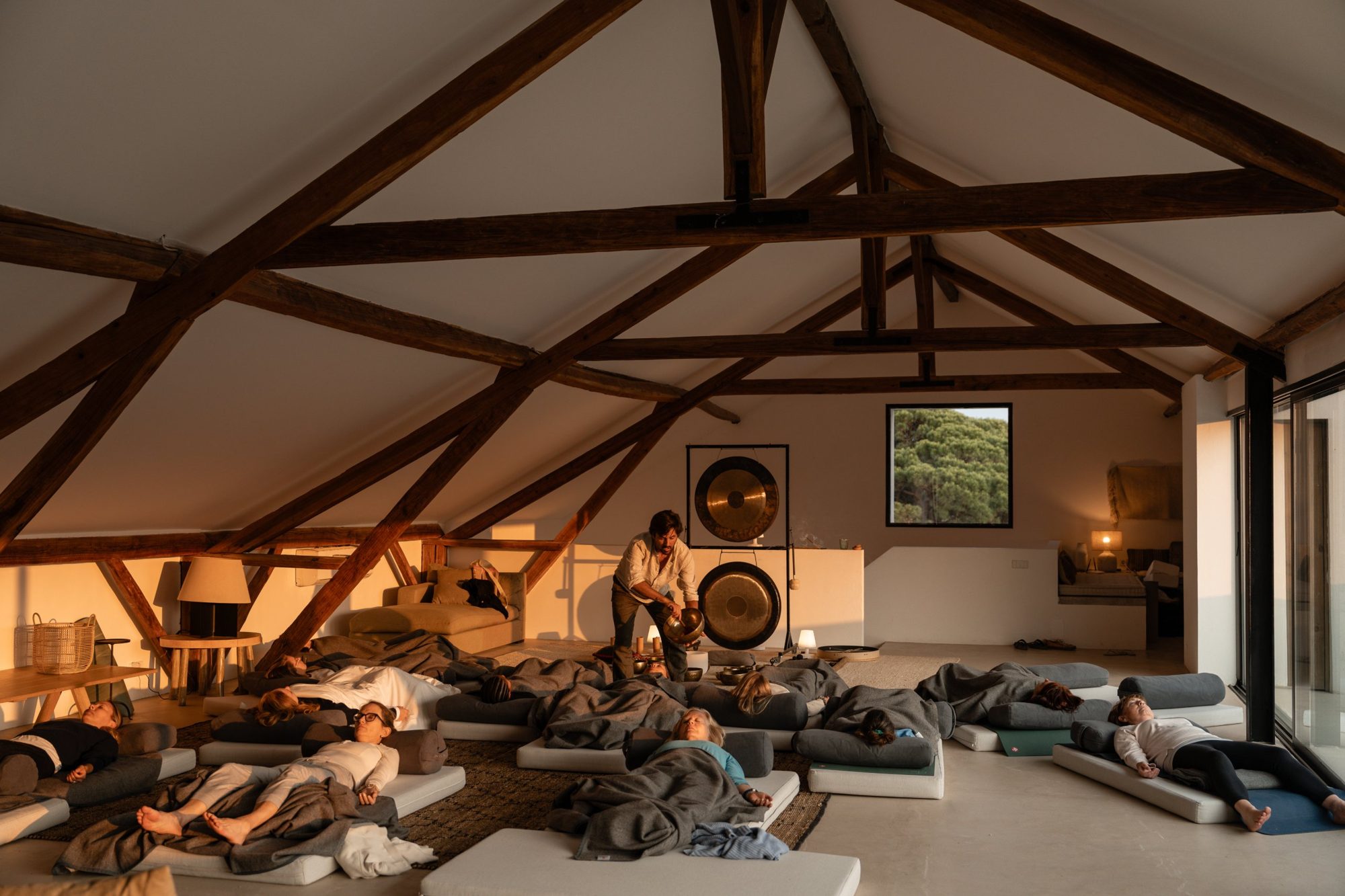 Francisco playing a singing bowl over a group resting on mattresses under wooden beams at sunset